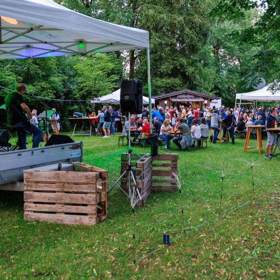 Eine lebhafte Outdoor-Veranstaltung mit Menschen an Tischen, einer Bühne mit einem Musiker und verschiedenen Zelten und Lautsprechern in einer parkähnlichen Umgebung.