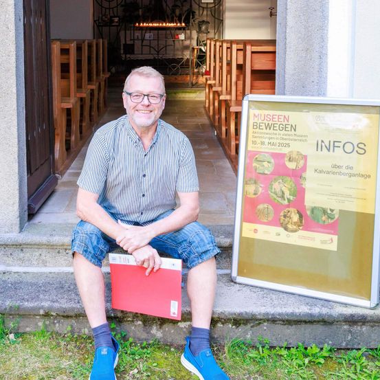 Ein Mann sitzt auf den Stufen einer Kirche, lächelt, hält eine rote Mappe in der Hand, mit einem Schild hinter ihm, das 'Museen bewegen' und 'Infos' besagt.
