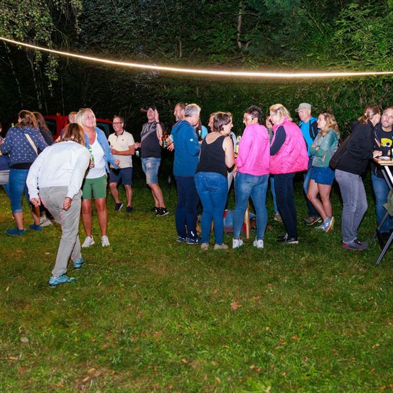 In der Nacht versammelt sich eine Gruppe von Menschen in einem Waldgebiet, steht und unterhält sich. Einige halten Flaschen, und ein Tisch mit Flaschen befindet sich in der Nähe. Bäume umgeben das Gebiet.