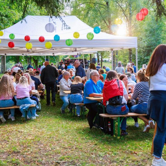 Eine große Menschenmenge versammelt sich unter einem weißen Zelt mit bunten Luftballons. Mehrere Personen sitzen an Tischen, andere stehen herum, einige mit Getränken. Bäume und Grün umgeben den Bereich.