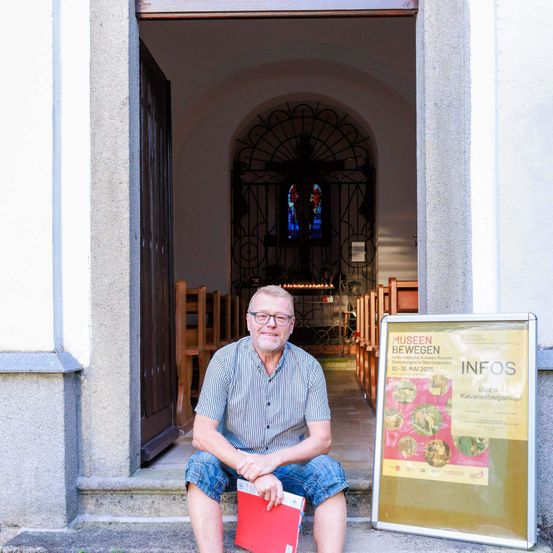 Ein Mann mit Brille und gestreiftem Hemd sitzt auf den Stufen einer Kirche mit einem roten Ordner im Schoß. Hinter ihm ist das Kircheninnere zu sehen, einschließlich einer Holztür, Kirchenbänken und einem kunstvollen eisernen Tor. Ein Informationsschild mit dem Wort 'Infos' ist an der Wand montiert.