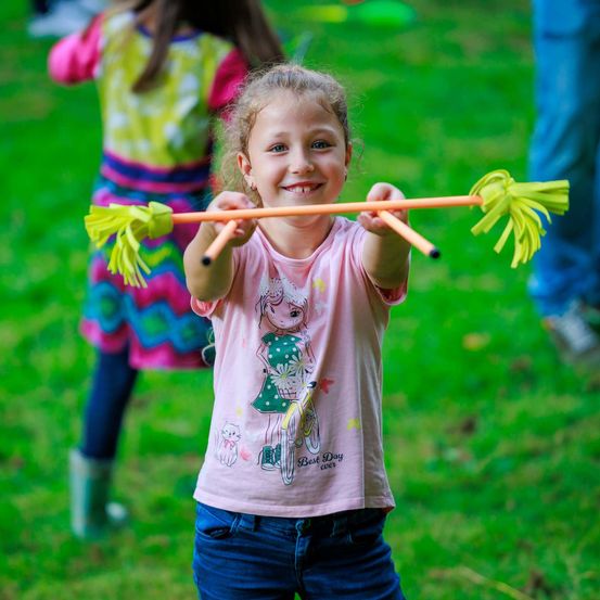 Ein kleines Mädchen hält zwei Stöcke mit gelben Pompons und lächelt. Sie trägt ein rosa T-Shirt und blaue Jeans. Hinter ihr spielt ein anderes Kind mit einem Frisbee.