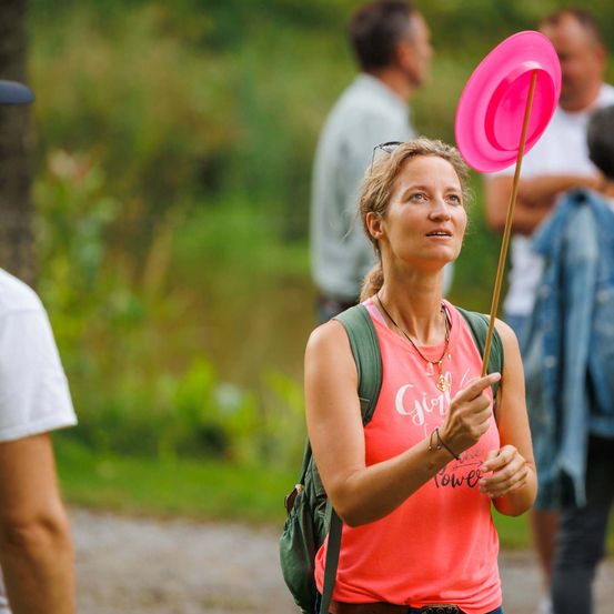 Eine Frau mit Rucksack und pinkem Frisbee steht in einer Outdoor-Umgebung, umgeben von verschwommenen Personen.