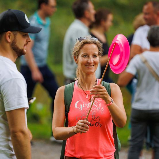 Eine Frau, die ein rosa Frisbee hält, lächelt, während sie unter mehreren Personen steht, wahrscheinlich bei einer Veranstaltung im Freien.