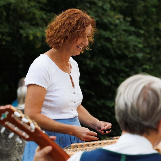 Eine ältere Frau mit lockigem rotem Haar und einem weißen Oberteil lächelt, während sie mit einem Mann mit grauem Haar eine Zither spielt. Sie sind draußen in der Nähe einer Steinmauer.