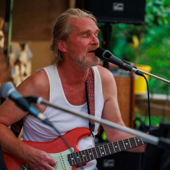Ein Mann spielt eine E-Gitarre in ein Mikrofon, trägt ein weißes Tanktop und einen roten Gurt. Er hat die Augen geschlossen und ist von üppiger grüner Vegetation umgeben.