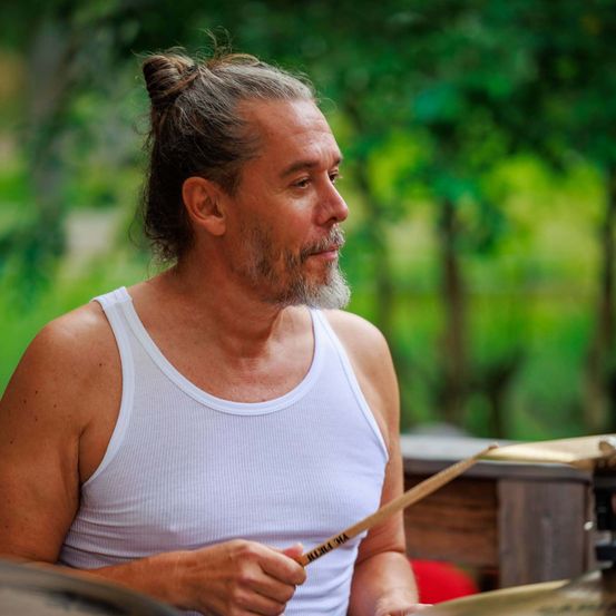 Ein bärtiger Mann spielt draußen Schlagzeug, gekleidet in einem weißen Tanktop, unter einem sonnigen Himmel.
