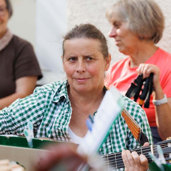 Eine Frau mit kurzen Haaren spielt Gitarre und lächelt in die Kamera. Hinter ihr hält eine andere Frau in einem roten T-Shirt schwarze Handschuhe. Eine weitere Frau im Hintergrund trägt eine Brille und ein braunes T-Shirt.