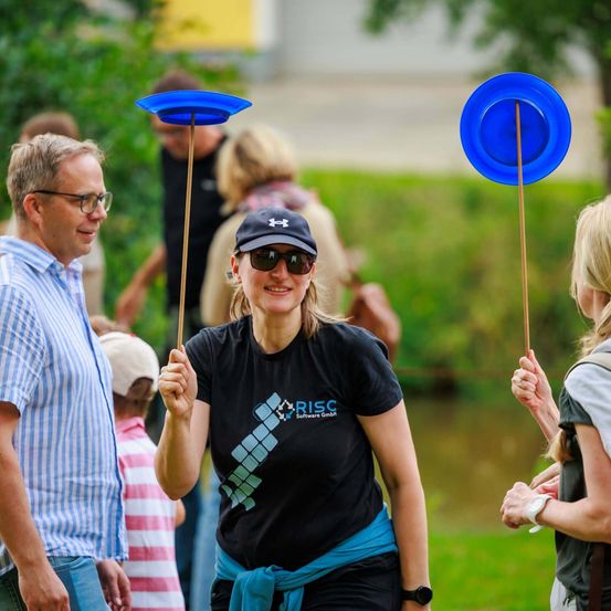 Eine Frau mit einem schwarzen T-Shirt, auf dem RISC Software GmbH steht, hält draußen einen blauen Teller auf einem Stock. In der Nähe stehen ein Mann und ein Kind hinter ihr, und eine Frau trägt ein Kind auf der Schulter.
