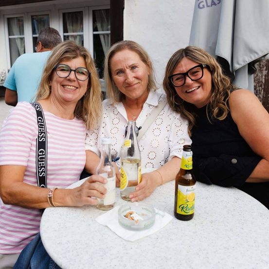 Drei Frauen mit Brille sitzen bei Getränken am Tisch und lächeln in die Kamera. Hinter ihnen ist ein Mann teilweise zu sehen.