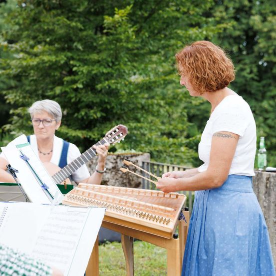 Zwei Frauen spielen Instrumente im Freien. Eine spielt eine Harfe und die andere eine Gitarre. Sie tragen beide eine Brille und sehen sich Noten an.