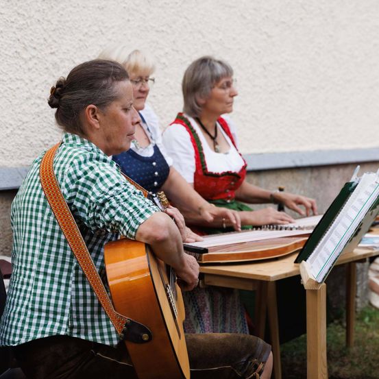 Drei Frauen sitzen vor einer Wand, eine spielt Gitarre und die anderen beiden spielen Zither. Sie tragen traditionelle Kleidung.