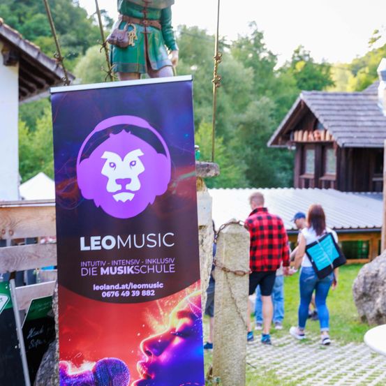 Ein Banner einer Musikschule mit einem Löwenlogo und der Website-Adresse steht vor einem Gebäude. Ein Mann und eine Frau gehen Hand in Hand, mit Bäumen und einem weiteren Gebäude im Hintergrund.