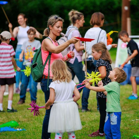 Eine Gruppe von Kindern und Erwachsenen befindet sich auf einem Grasfeld. Einige halten Spielzeug, und eine Frau hält eine rote Frisbee. Hinter ihnen sind Bäume und ein Pfahl.