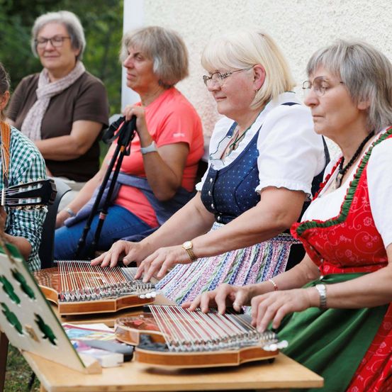 Fünf ältere Frauen spielen traditionelle Saiteninstrumente im Freien. Sie sind in traditioneller Kleidung gekleidet. Eine von ihnen spielt eine Zither, während eine andere ein anderes Saiteninstrument spielt.