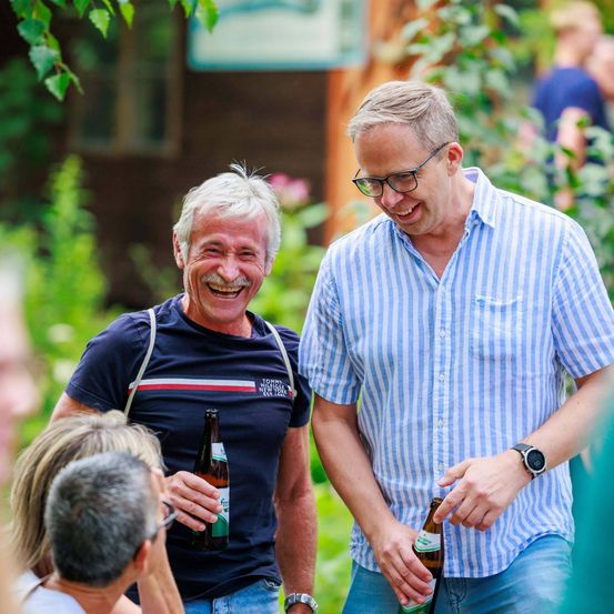 Zwei Männer in einem Garten, lächelnd, einer hält eine Bierflasche. Eine Frau mit blonden Haaren ist in der Nähe, und ein Holzhaus mit einem Fenster ist im Hintergrund.
