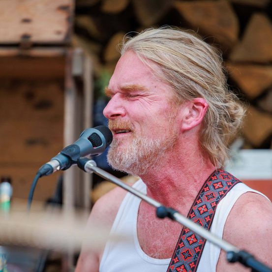 Ein Mann mit langen Haaren und Bart singt in ein Mikrofon. Er trägt ein weißes Tanktop und einen bunten Gurt über der Schulter. Dahinter steht eine Holzwand mit Holzscheiten.