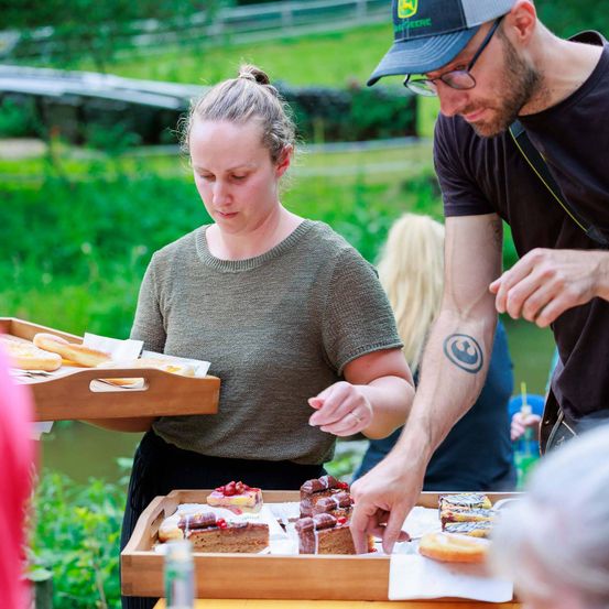 Eine Frau serviert Gebäck auf einem Tablett, während ein Mann mit Brille und Kappe ein Gebäck auswählt. Sie befinden sich in einer Outdoor-Umgebung mit Grünflächen und anderen Menschen.