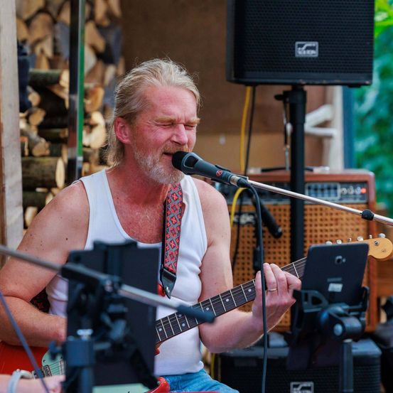 Ein Mann mit Bart und Schnurrbart spielt Gitarre und singt in ein Mikrofon. Er trägt ein weißes Tanktop und einen Gitarrengurt. Hinter ihm steht ein Lautsprecher und weitere Ausrüstung.
