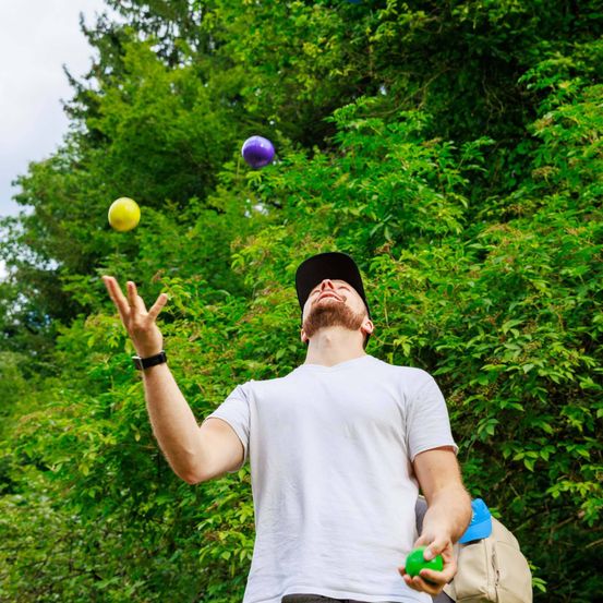Ein Mann jongliert mit drei bunten Bällen im Freien in einer üppigen grünen Umgebung. Er trägt ein weißes T-Shirt und eine schwarze Kappe.