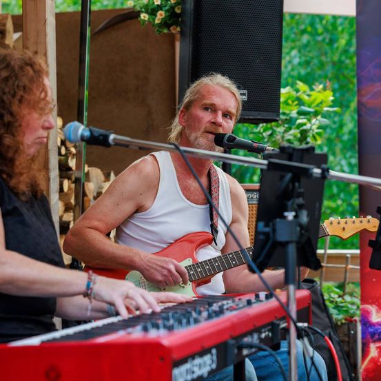 Ein Mann spielt eine rote Gitarre, während er neben einer Frau sitzt, die eine Tastatur spielt. Sie singen beide in Mikrofone.