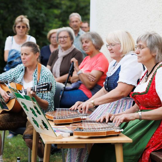 Eine Gruppe älterer Frauen sitzt im Freien auf Stühlen und spielt Instrumente. Eine Frau spielt Gitarre, eine andere spielt Zither. Sie tragen traditionelle Kleidung. Hinter ihnen sitzen mehrere andere Frauen und beobachten.