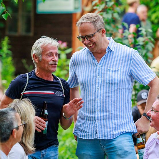 Zwei Männer lächeln und lachen, während sie im Freien stehen, einer in einem Tommy Hilfiger-Shirt und der andere in einem blau-weiß gestreiften Hemd. Hinter ihnen versammelt sich eine Gruppe von Menschen in einem Garten.