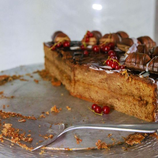 Ein Stück Kuchen mit Schokoladenglasur und roten Beeren liegt auf einem Teller mit einem Gabel in der Nähe, Krümel verstreut.