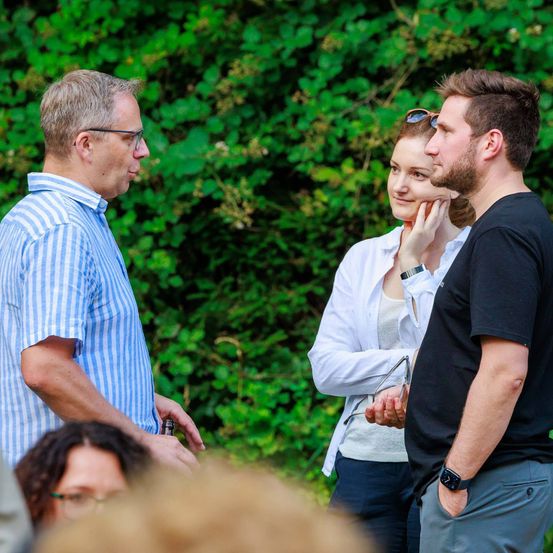 Drei Personen stehen in einer Outdoor-Umgebung, wobei ein Mann in einem blau gestreiften Hemd mit einem Mann in einem schwarzen T-Shirt spricht. Eine Frau in einer weißen Jacke mit einer Brille auf dem Kopf lächelt. Hinter ihnen ist eine dichte Begrünung.