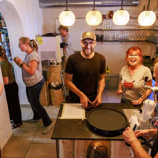 Eine Gruppe von Menschen ist in einem Café. Ein Mann steht hinter der Theke mit einer Frau auf seiner rechten und einer anderen Frau auf seiner linken Seite. Es gibt Flaschen und Tassen in den Regalen hinter ihnen.