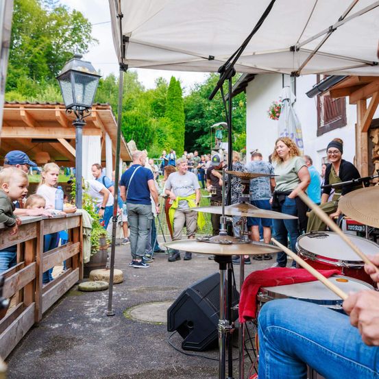 Eine Gruppe von Menschen beobachtet eine Band, die auf einer Bühne unter einem Zelt spielt. Der Schlagzeuger sitzt rechts, während die Becken und Trommeln vor ihm aufgestellt sind. In der Nähe stehen Menschen und beobachten.