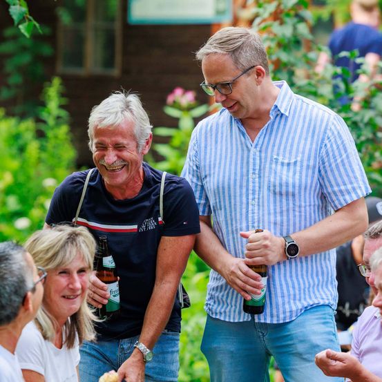 Zwei Männer und eine Frau stehen in einem Garten, lächeln und halten Flaschen. Ein Mann trägt eine Uhr, der andere ein gestreiftes Hemd. Eine weitere Frau mit blonden Haaren lächelt sie an.