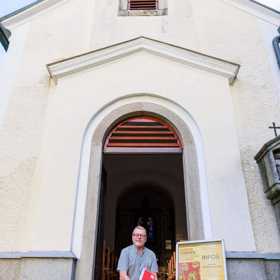 Ein Mann steht vor einer Kirche und hält ein Buch in der Hand. Die Kirche hat einen gewölbten Eingang und ein Schild mit 'MUSEUM' und 'INFOS' darauf.