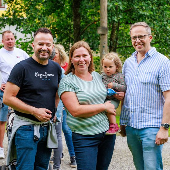 Eine Familie mit drei Personen, darunter ein Baby, steht vor einem Baum. Der Mann links trägt ein Pepe Jeans T-Shirt. Die Frau lächelt und hält das Baby. Der Mann rechts trägt eine Brille und eine Uhr. Im Hintergrund sind weitere Personen zu sehen.