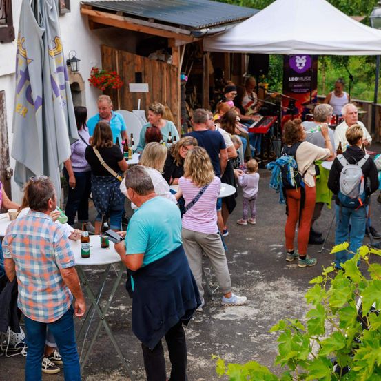 Mehrere Menschen versammeln sich draußen in einer Café-Atmosphäre. Einige stehen und unterhalten sich, andere sitzen an Tischen. Zelte und ein Musikständer sind aufgestellt.