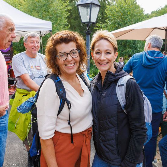 Zwei Frauen stehen bei einer Outdoor-Veranstaltung und lächeln. Eine trägt eine Brille, ein weißes Oberteil und orangefarbene Hosen und hat einen Rucksack dabei. Die andere hat kurze Haare und trägt eine schwarze Jacke. Hinter ihnen versammeln sich Leute unter Schirmen und Zelten.