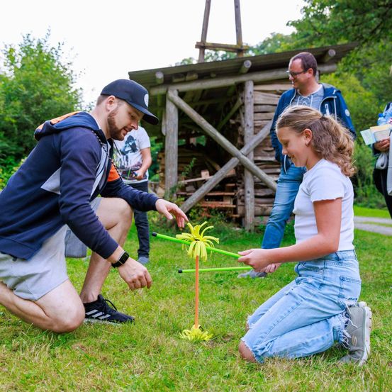 Ein Mann und ein Mädchen spielen mit einem gelben Blumenspielzeug auf dem Rasen, in der Nähe einer Holzstruktur mit Brennholz und einem Mann mit Brille, der beobachtet.