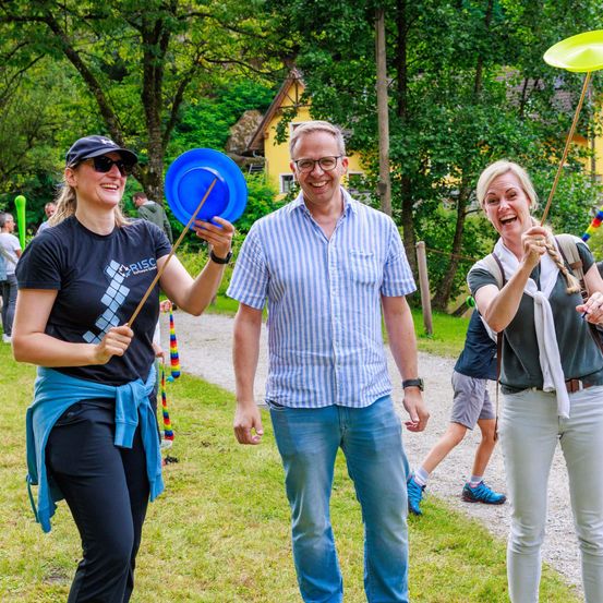 Drei Erwachsene lächeln und lachen in einem Park und halten bunte Stöcke und Teller. Ein gelbes Haus ist im Hintergrund.