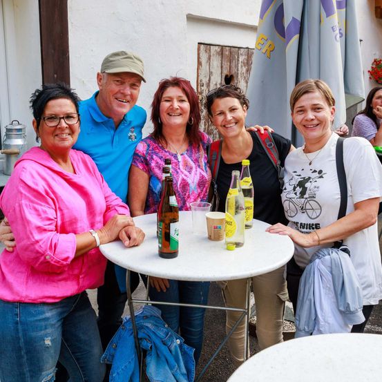 Eine Gruppe von fünf Erwachsenen steht um einen Tisch mit Getränken und lächelt für ein Foto. Hinter ihnen ist ein Gebäude und Sonnenschirme zu sehen.
