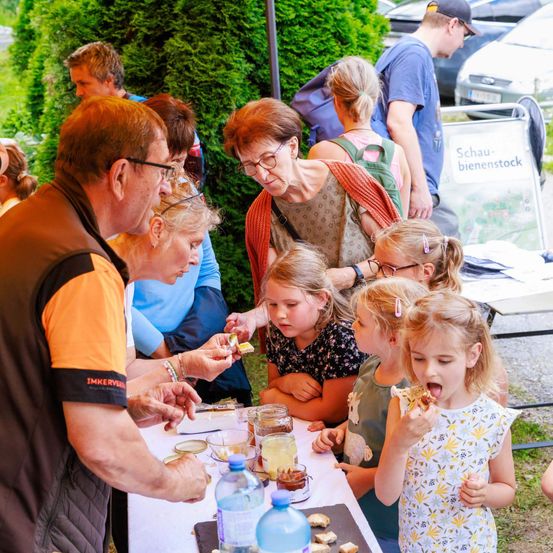 Eine Gruppe von Erwachsenen und Kindern versammelt sich um einen Tisch mit Essen. Ein erwachsener Mann in einem braunen Hemd spricht mit den Kindern. Die Kinder essen und genießen das Essen. Hinter ihnen steht ein Schild mit Text und ein geparktes Auto.