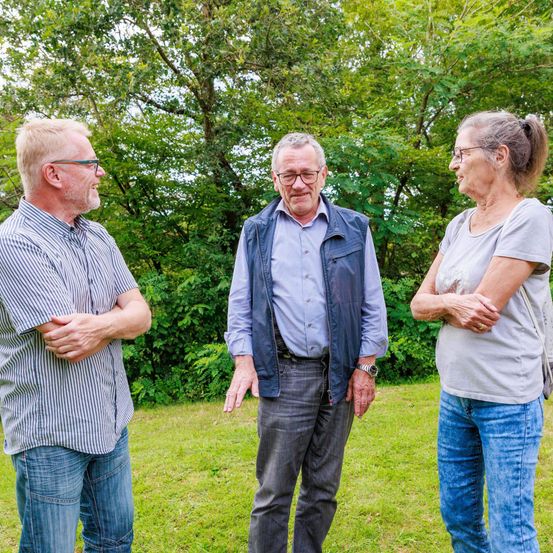 Drei Menschen stehen in einem Grasbereich, wahrscheinlich in einem Park. Sie alle lächeln und scheinen zu plaudern. Der Mann auf der linken Seite trägt eine Brille und hat die Arme verschränkt. Die mittlere Person trägt eine Weste und eine Uhr. Die Frau auf der rechten Seite trägt ein graues Shirt und hat die Arme gefaltet. Bäume stehen hinter ihnen.