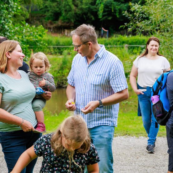 Eine Familie geht entlang eines Kiesweges in der Nähe eines Flusses. Ein Mann hält etwas in der Hand, und eine Frau mit einem Baby schaut ihn an. Ein Mädchen schaut nach unten, und eine weitere Frau ist hinter ihnen.