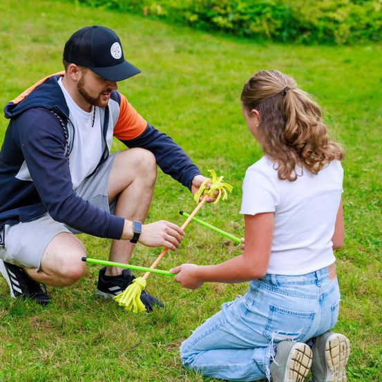 Ein Mann und ein Mädchen knien auf dem Gras und halten grüne und gelbe Objekte. Der Mann trägt eine Kappe und Turnschuhe, während das Mädchen eine Pferdeschwanzfrisur hat und Stiefel trägt.