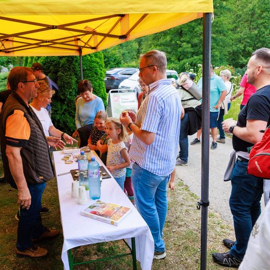 Mehrere Personen versammeln sich unter einem gelben Zelt, möglicherweise bei einer Gemeinschaftsveranstaltung. Ein Mann spricht zu einer Gruppe, und ein Tisch mit Büchern und Flaschen ist aufgebaut. Bäume und geparkte Autos sind im Hintergrund zu sehen.