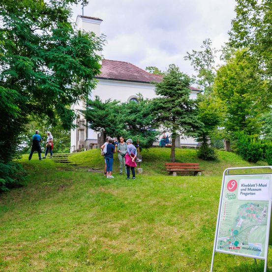 Eine Gruppe von Menschen besucht Kleeblatt-1-Musi und Museum Pregarten, umgeben von Bäumen und Gras. Eine Bank steht auf dem Rasen.
