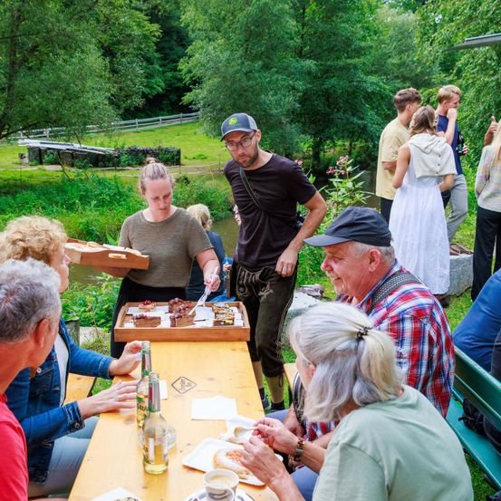 Eine Gruppe von Menschen sitzt um einen Holztisch im Freien. Eine Frau serviert Essen von einem Tablett, während ein Mann davor lächelt. Dahinter stehen Bäume und ein kleiner Bach.