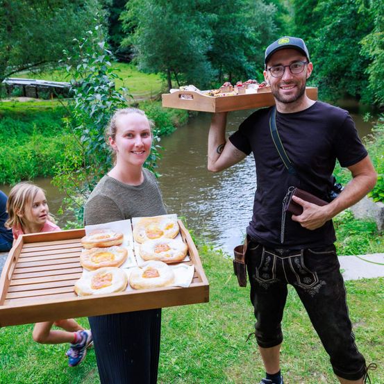 Ein Mann und eine Frau halten einen Tablett mit Essen in einem Grasbereich in der Nähe eines Flusses. Der Mann trägt ein schwarzes Shirt und Hut, während die Frau in einem grauen Oberteil ist. Hinter ihnen geht ein junges Mädchen in der Nähe des Flusses.