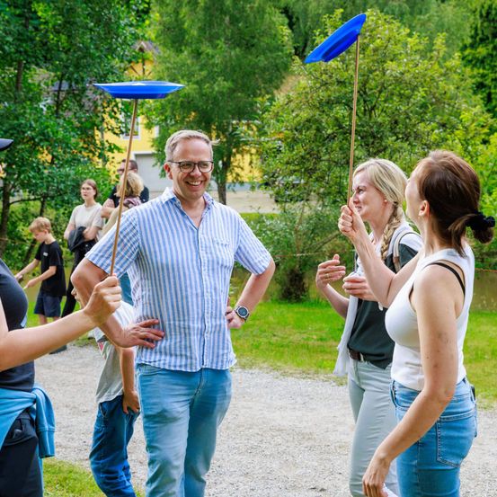 Eine Gruppe von Menschen steht in einem Park, ein Mann in einem blau-weiß gestreiften Hemd lächelt. Zwei Frauen halten blaue Frisbees. Hinter ihnen spricht eine Frau mit einem Kind. Bäume und ein gelbes Gebäude sind im Hintergrund.