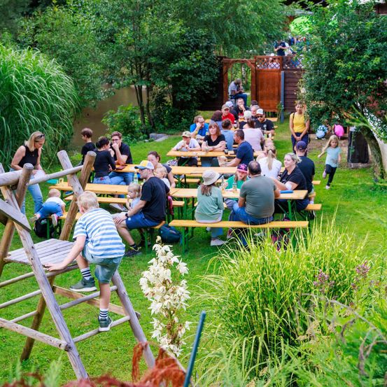 Ein Familientreffen in einem Garten mit einem Fluss im Hintergrund. Die Leute sitzen an Tischen und plaudern. Ein Kind klettert auf eine Holzleiter.