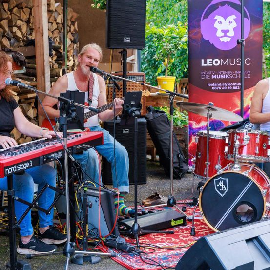 Eine Band spielt auf einer Bühne mit rotem Teppich. Ein Mann spielt Gitarre und singt in ein Mikrofon. Eine Frau spielt Keyboard und singt. Ein Schlagzeuger spielt das Schlagzeug. Es gibt ein Plakat mit dem Text 'LEOMUSIC'.